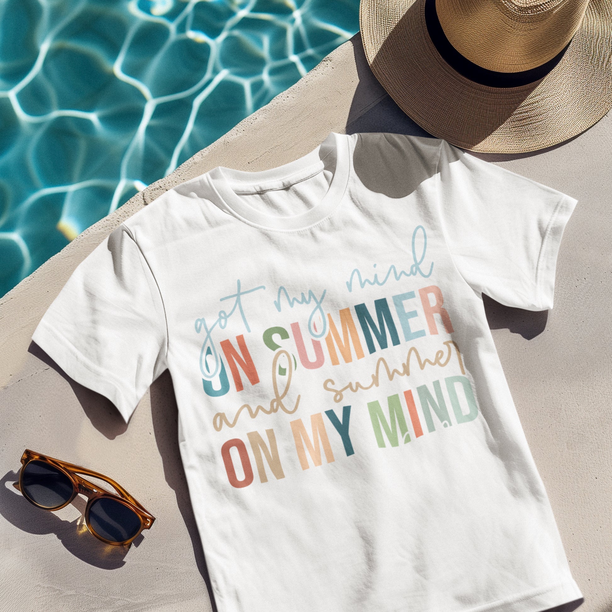 White summer quote t-shirt reading "Got My Mind on Summer and Summer on My Mind," laid next to sunglasses by the poolside. 