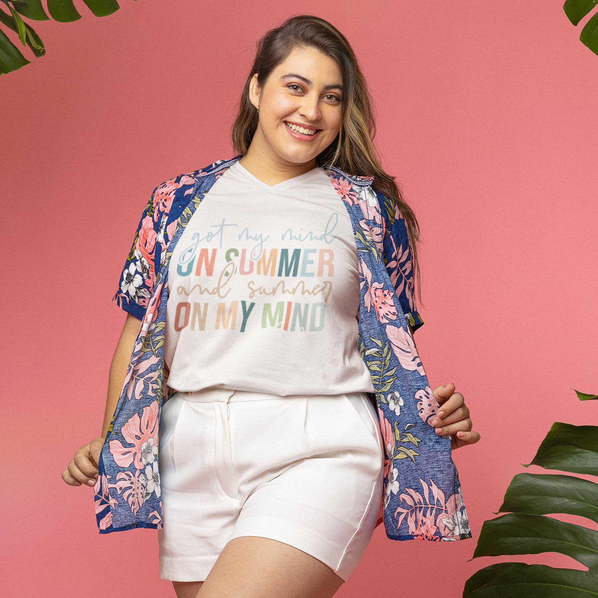 Woman wearing a summer quote t-shirt styled with a floral shirt and shorts, smiling against a pink background with tropical vibes. 