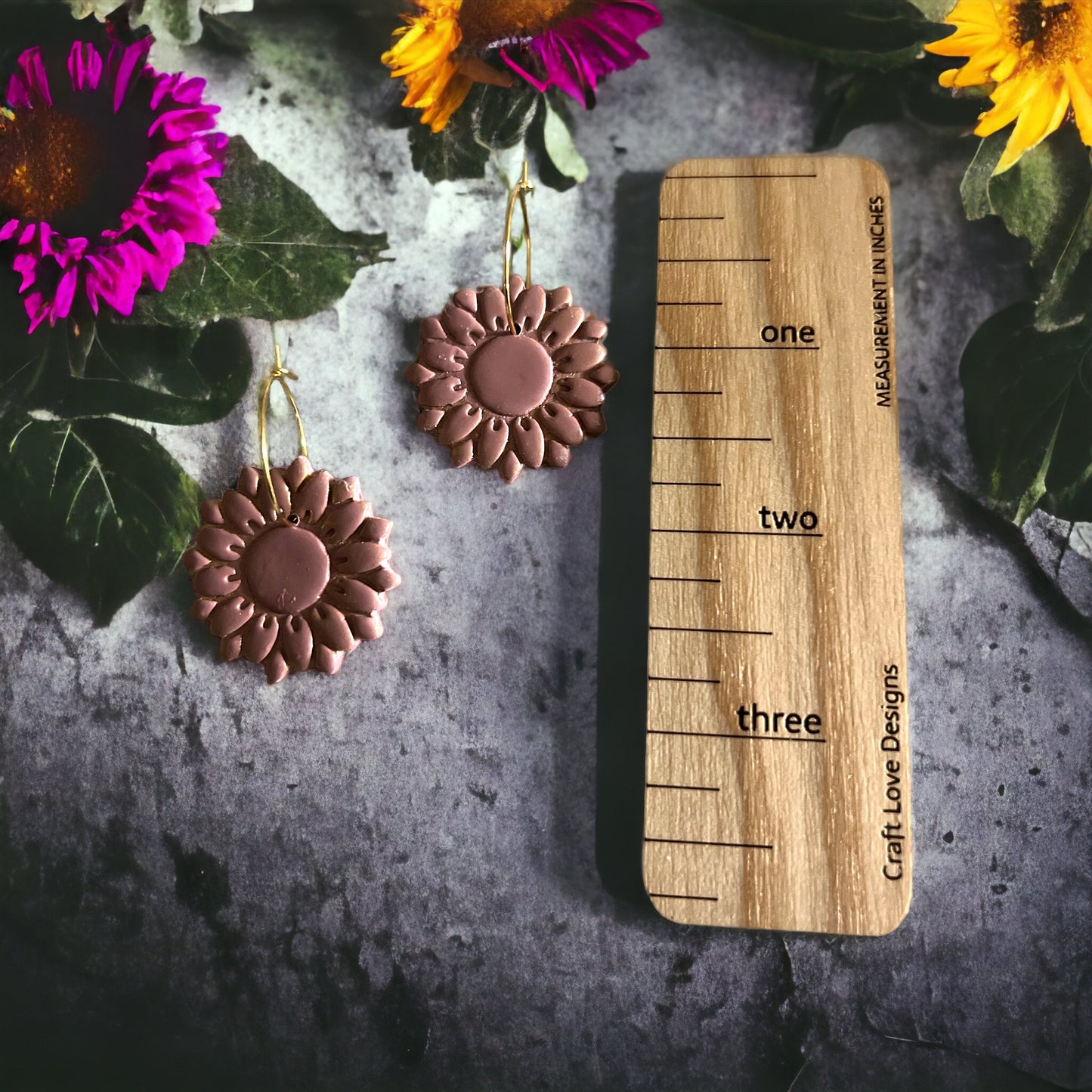 Dusty rose sunflower hoop earrings next to a measuring ruler, surrounded by colorful sunflowers on a rustic tabletop for size reference.