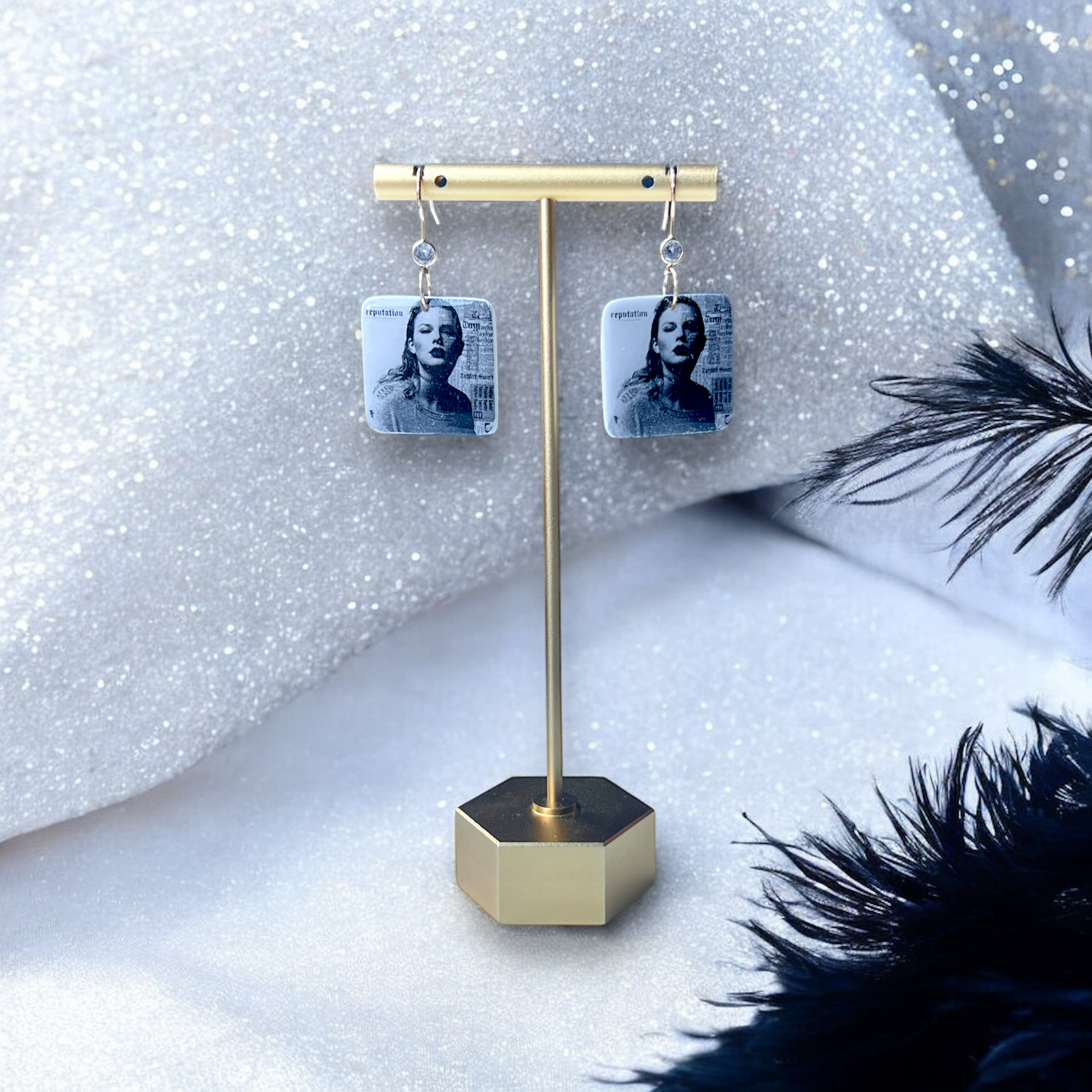 Reputation-inspired square earrings with black-and-white portrait details, displayed elegantly on a gold stand against a sparkly silver background.
