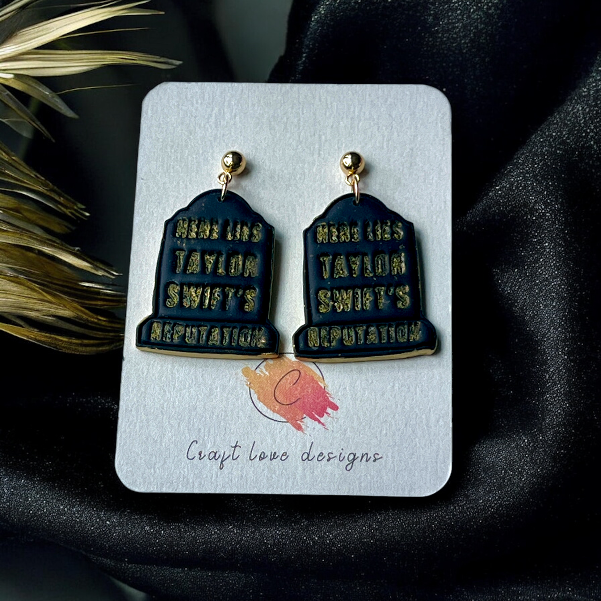 Black tombstone earrings with gold text reading "Here Lies Taylor Swift’s Reputation," displayed on a branded card against dark fabric. 