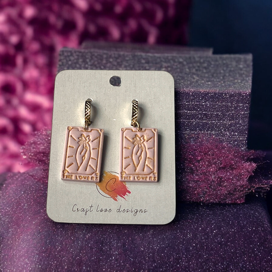Gold-accented dangle earrings featuring pastel pink tarot cards with embossed "The Lovers" design, displayed on a branded card. 