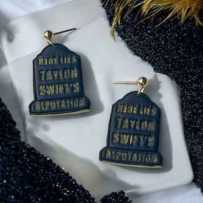 Black and gold tombstone-shaped earrings featuring embossed "Taylor Swift’s Reputation" text, resting on white fabric and sparkly black accents.