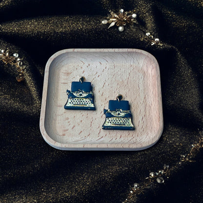Vintage-style black and gold typewriter earrings resting in a wooden tray, surrounded by sparkly fabric, pearls, and gold accents.