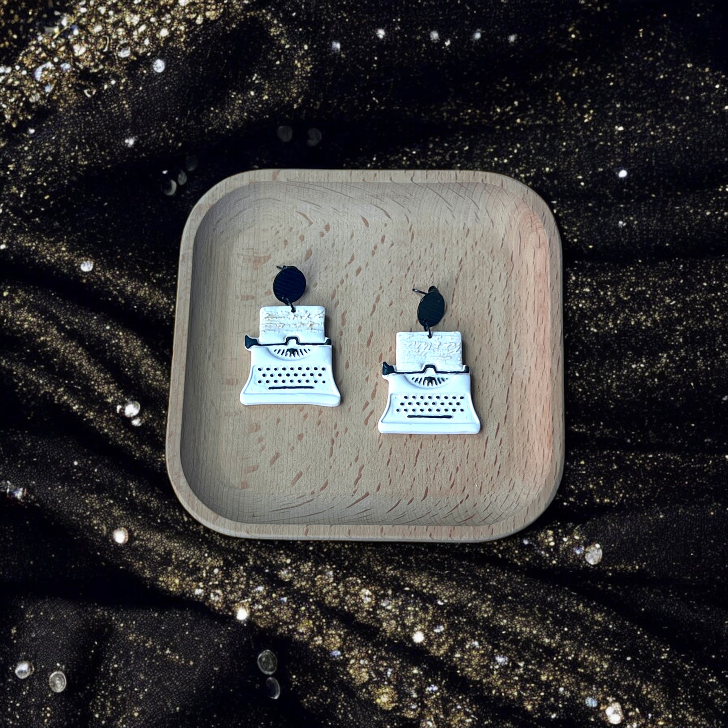 White and black typewriter earrings placed in a wooden tray, surrounded by glittering gold fabric and elegant pearl details.
(124 characters)