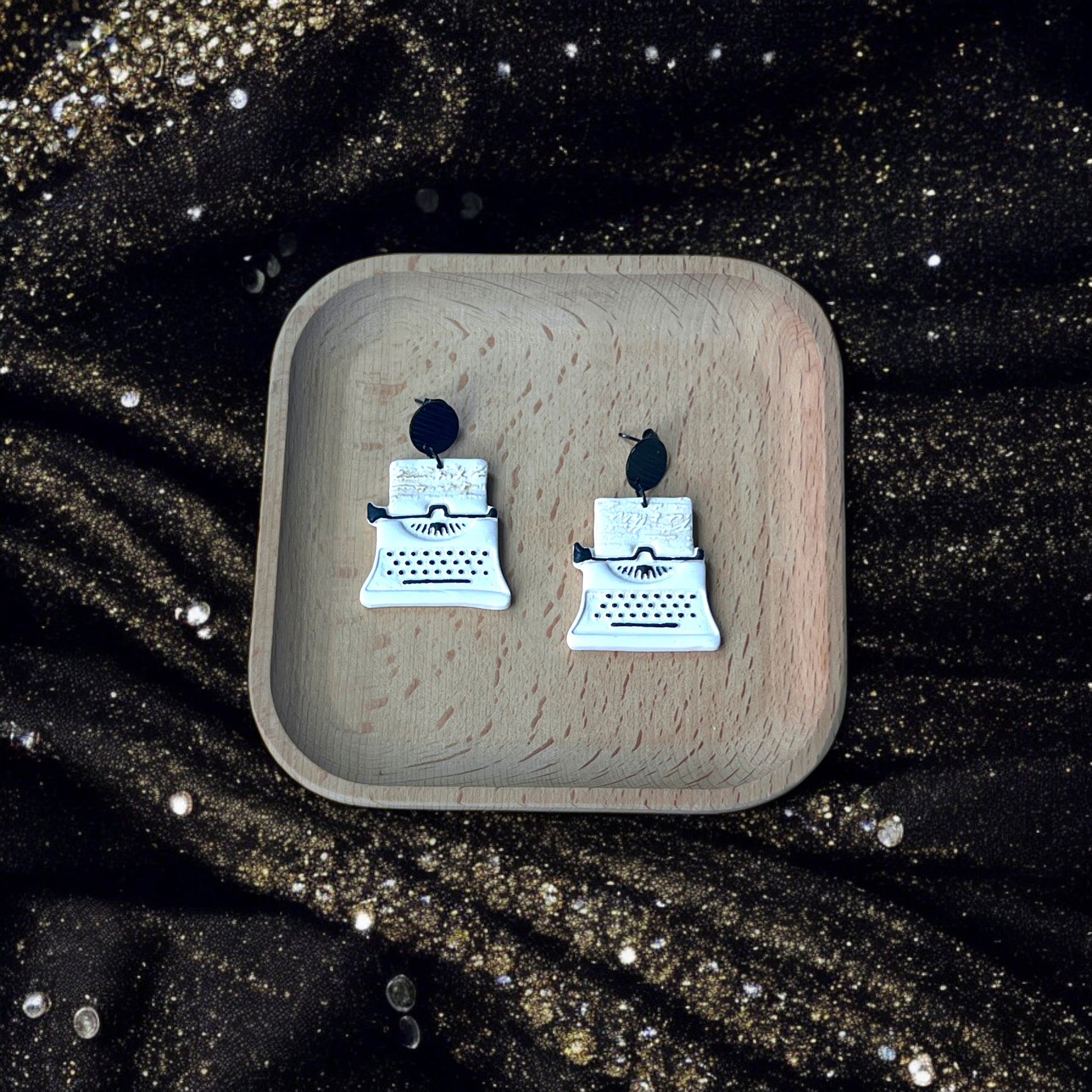 White and black typewriter earrings placed in a wooden tray, surrounded by glittering gold fabric and elegant pearl details.
(124 characters)
