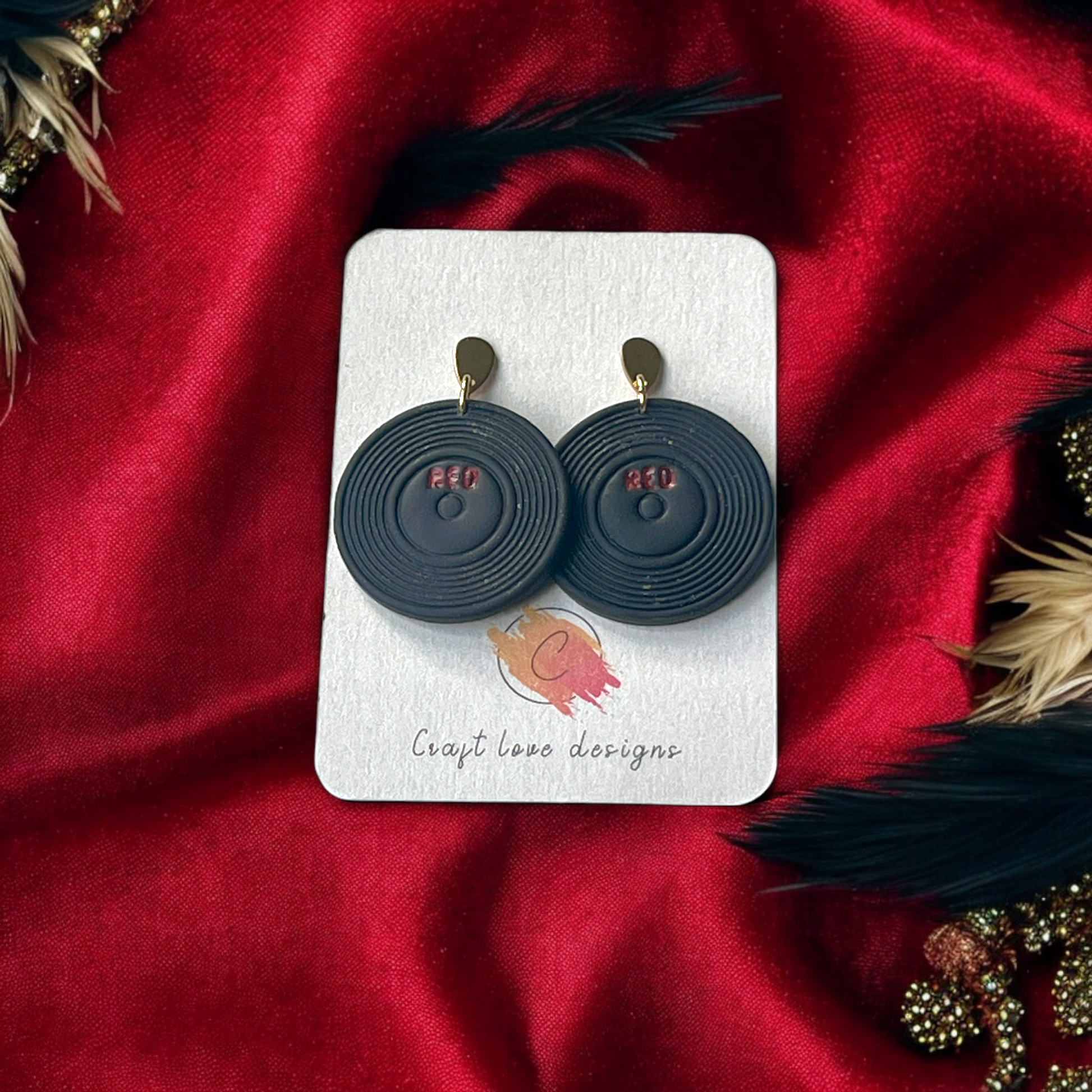 Black vinyl record-inspired dangle earrings with "Red" lettering, displayed on a Craft Love Designs card against a luxurious red fabric.