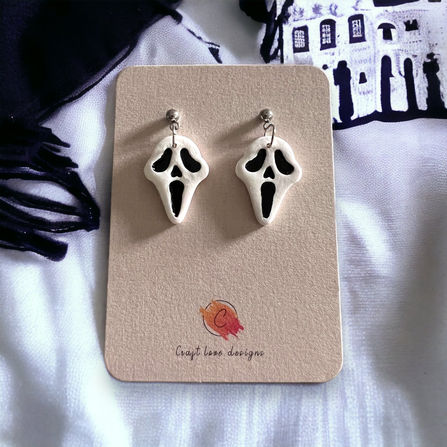 Close-up of white and black spooky face dangle earrings on a display card, surrounded by Halloween-themed decor like haunted house patterns.