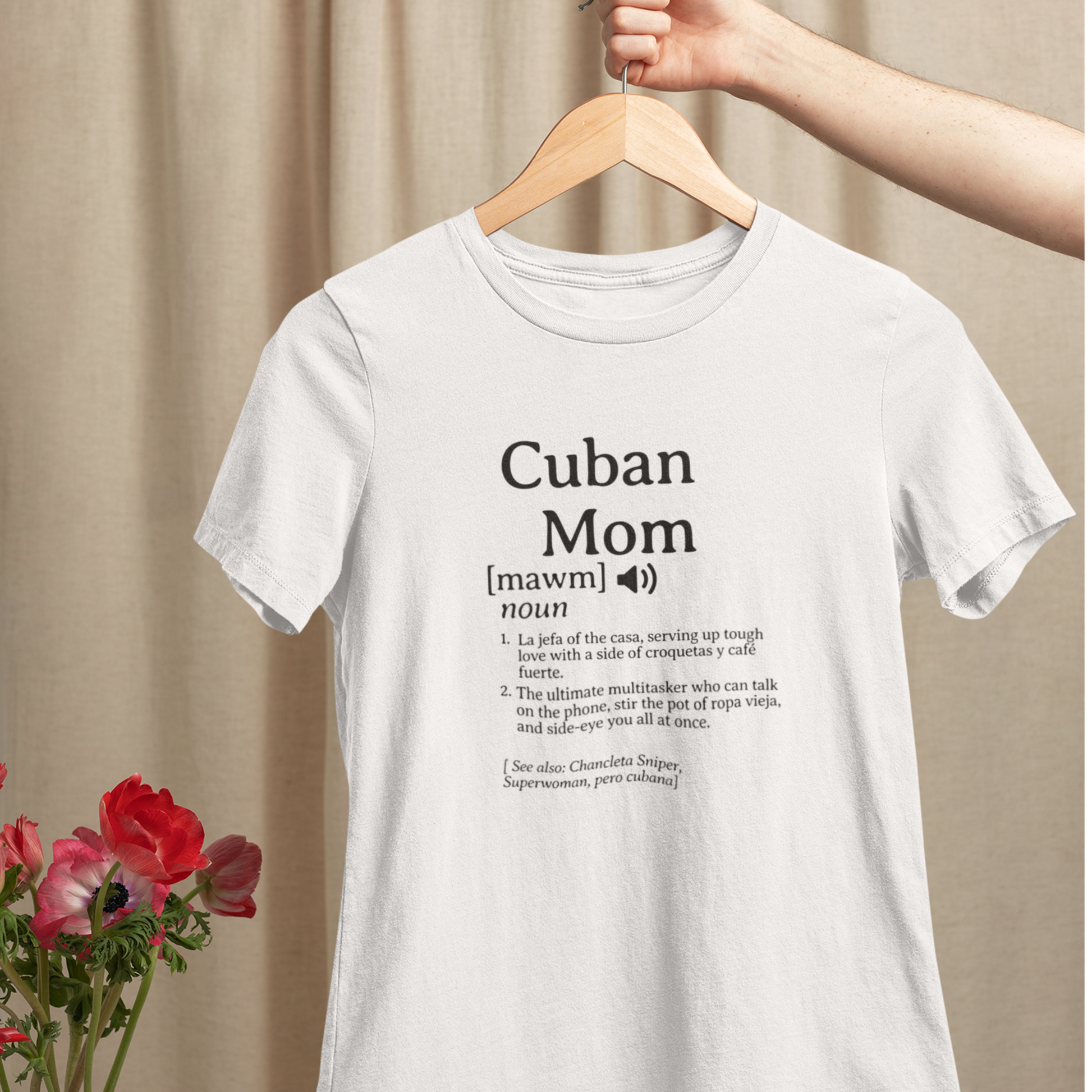 White Cuban Mom definition t-shirt hanging on a wooden hanger, displayed with nearby flowers against a soft neutral backdrop.background. 