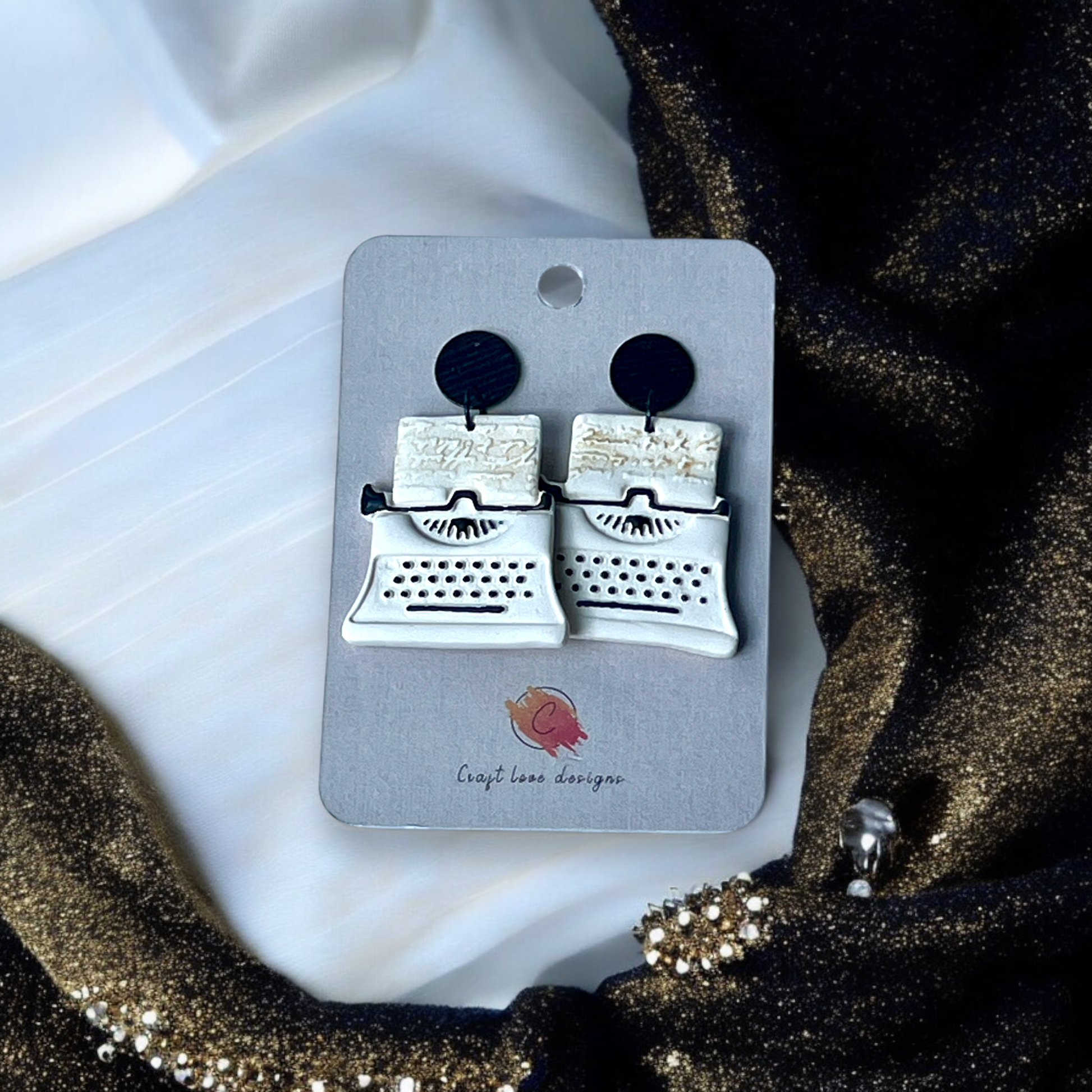 White and black vintage-inspired typewriter earrings displayed on a Craft Love Designs card, set against a gold and white fabric backdrop.