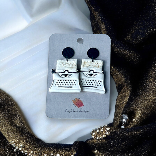 White and black vintage-inspired typewriter earrings displayed on a Craft Love Designs card, set against a gold and white fabric backdrop.