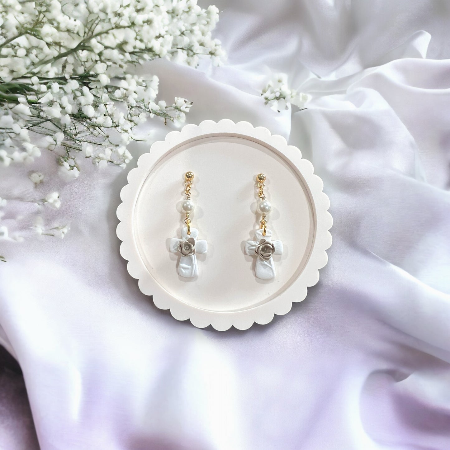 White pearl drop dangle earrings featuring a textured cross design with a delicate floral accent, displayed elegantly on a soft white fabric background.