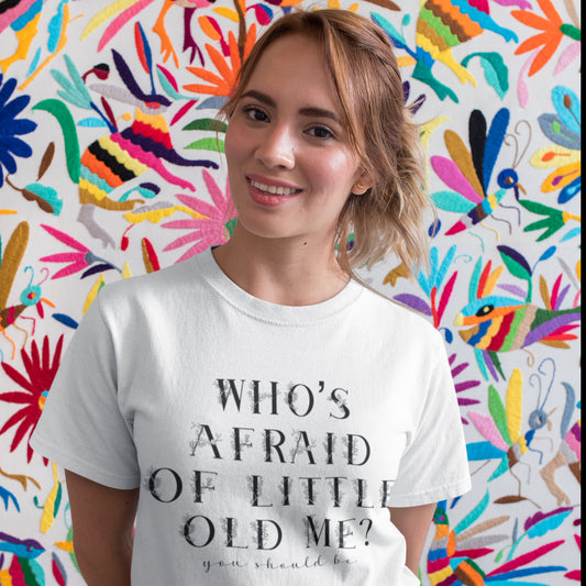 Woman wearing a white "Who’s Afraid of Little Old Me?" t-shirt, smiling in front of a vibrant and colorful background. 