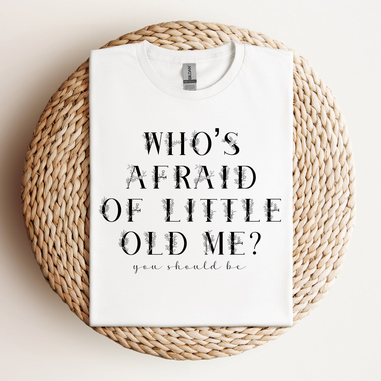 "Who’s Afraid of Little Old Me?" t-shirt neatly folded and displayed on a woven placemat with a minimal background.