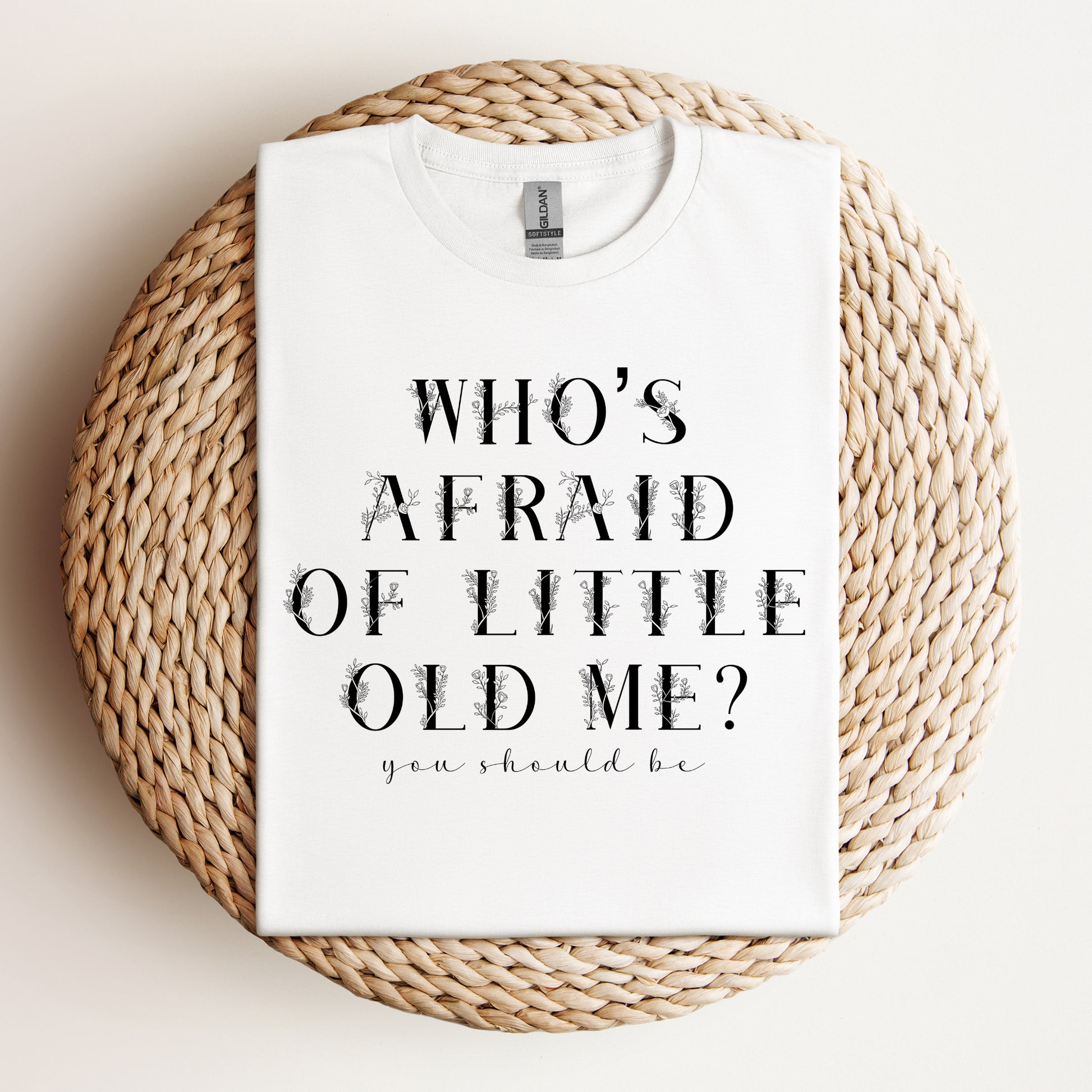 "Who’s Afraid of Little Old Me?" t-shirt neatly folded and displayed on a woven placemat with a minimal background.