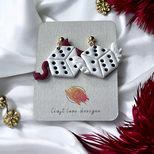Polymer clay statement earrings featuring white dice, one with devil horns and tail, and the other with angel wings, displayed on a branded card.