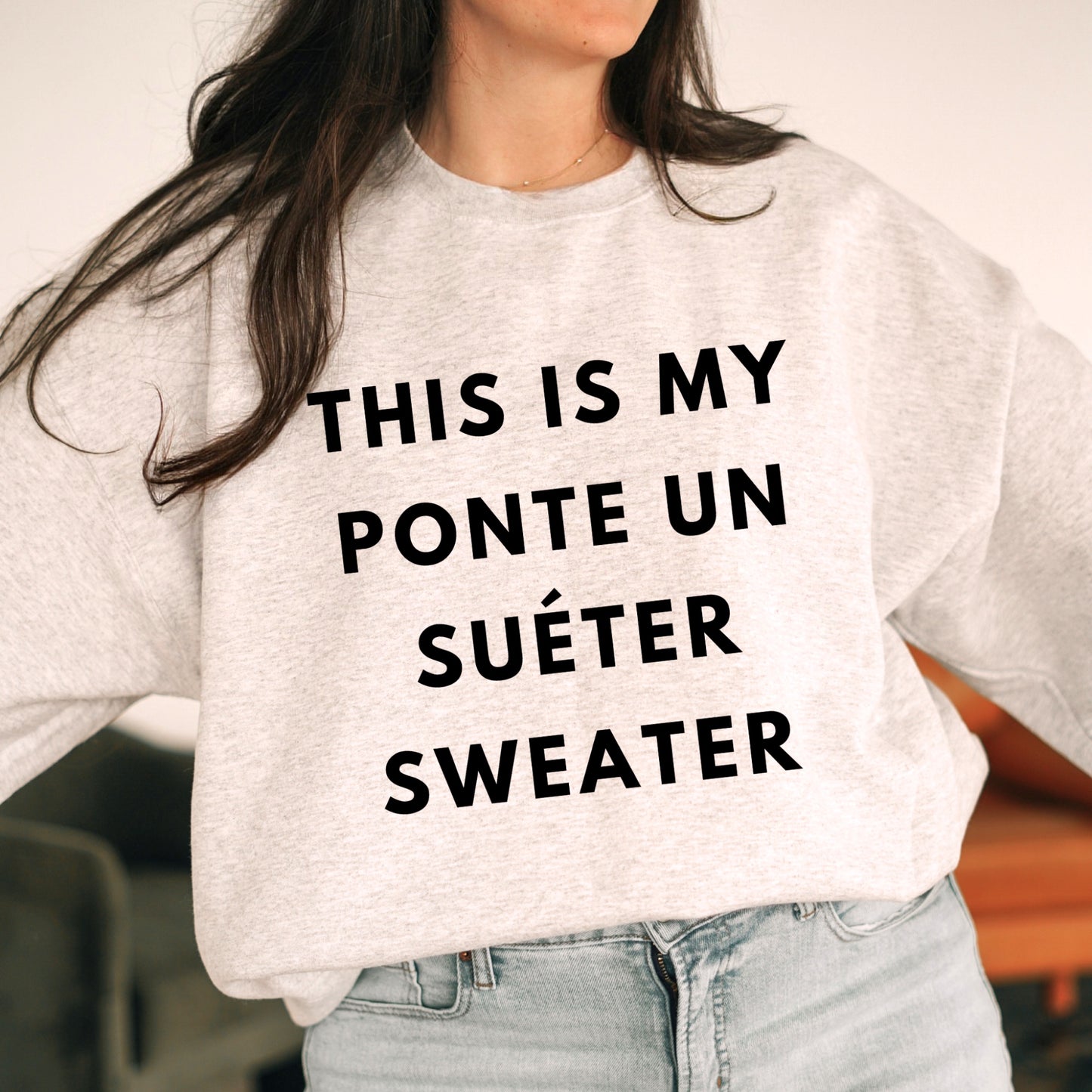 Woman wearing an ash gray "This Is My Ponte Un Suéter Sweater" crewneck with playful Spanish text, styled with light jeans indoors.