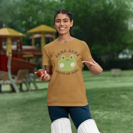Woman wearing mustard yellow t-shirt with a cute frog design and playful Spanish text "Sana Sana Colita de Rana," posing casually outdoors.