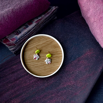 Handmade dangle earrings with yellow studs and floral charms displayed in a wooden tray, surrounded by soft pink and purple fabrics.