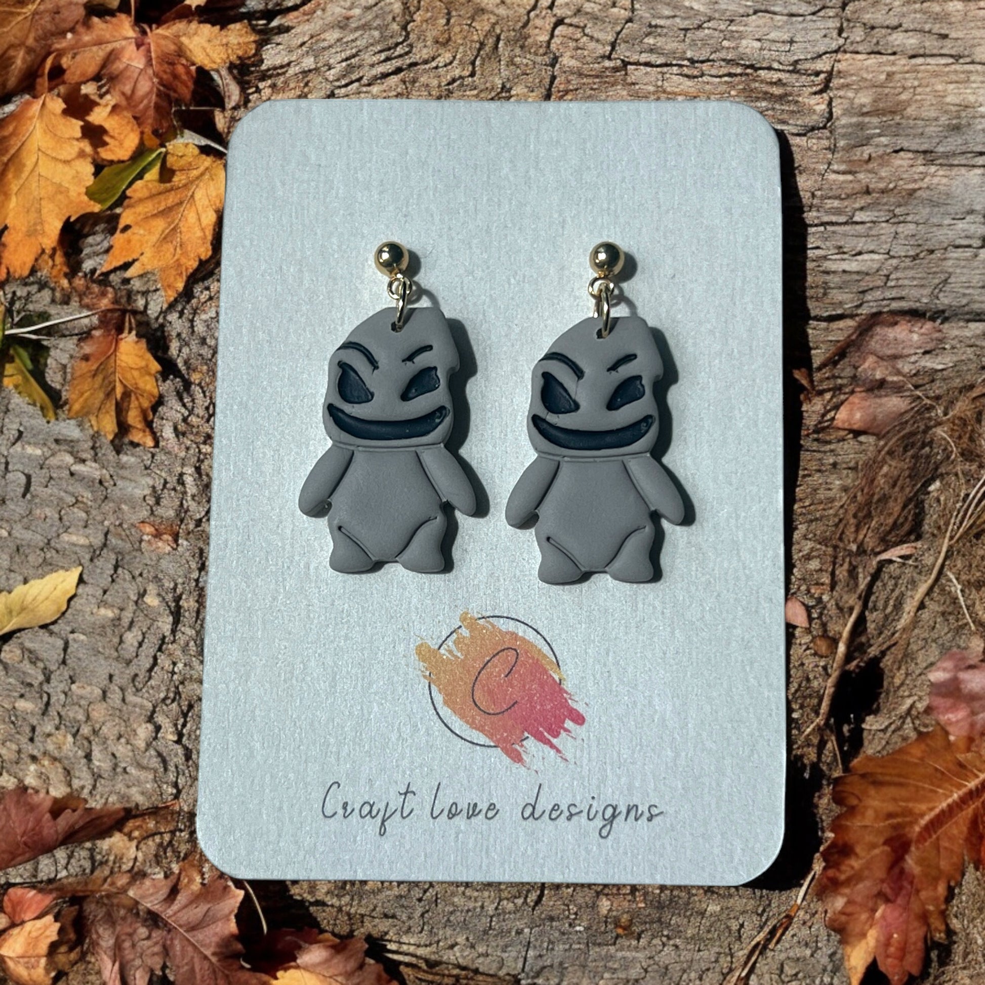 Woogie character earrings with mischievous grins on a Craft Love Designs display card, set against a rustic wooden background. 