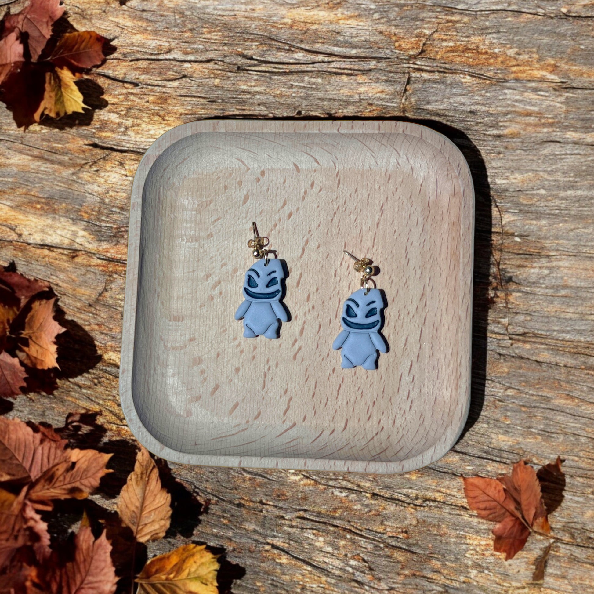 Woogie character earrings with smiling, mischievous faces resting in a wooden tray, surrounded by autumn leaves and rustic decor. 