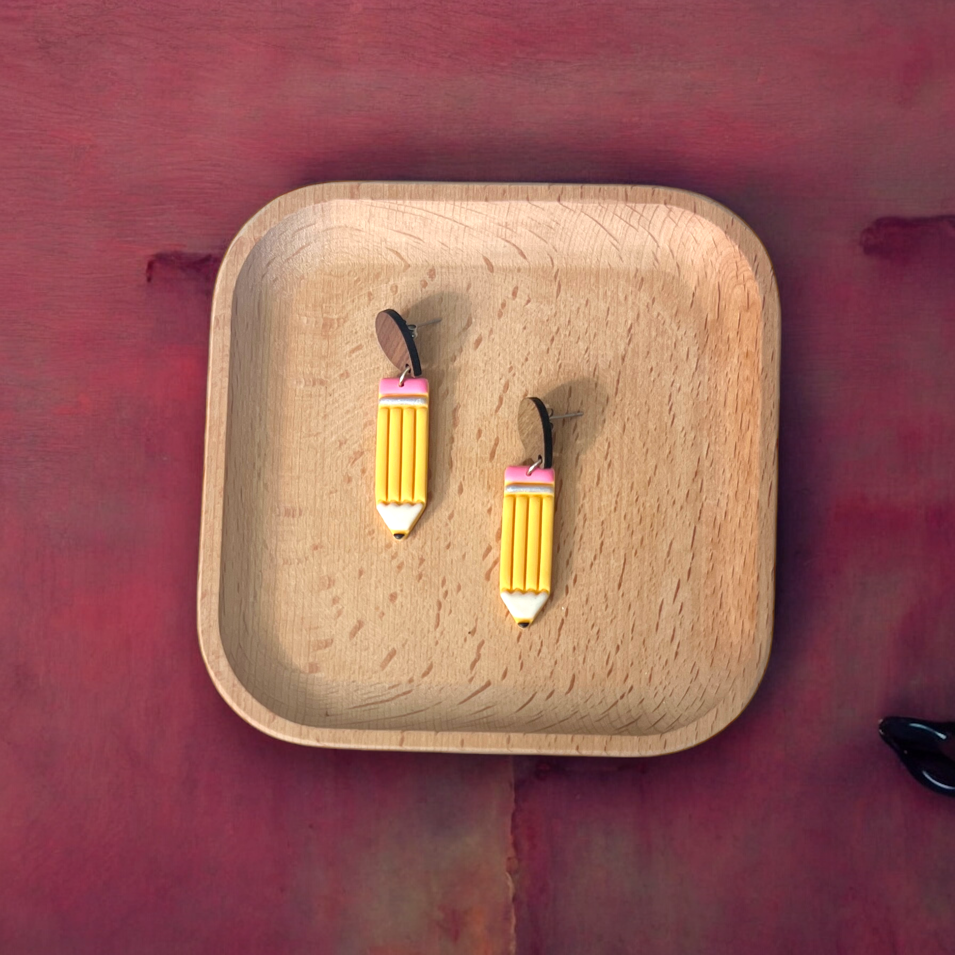 Cute pencil-themed drop earrings with yellow bodies and pink erasers, resting on a wooden tray against a soft pink surface. 