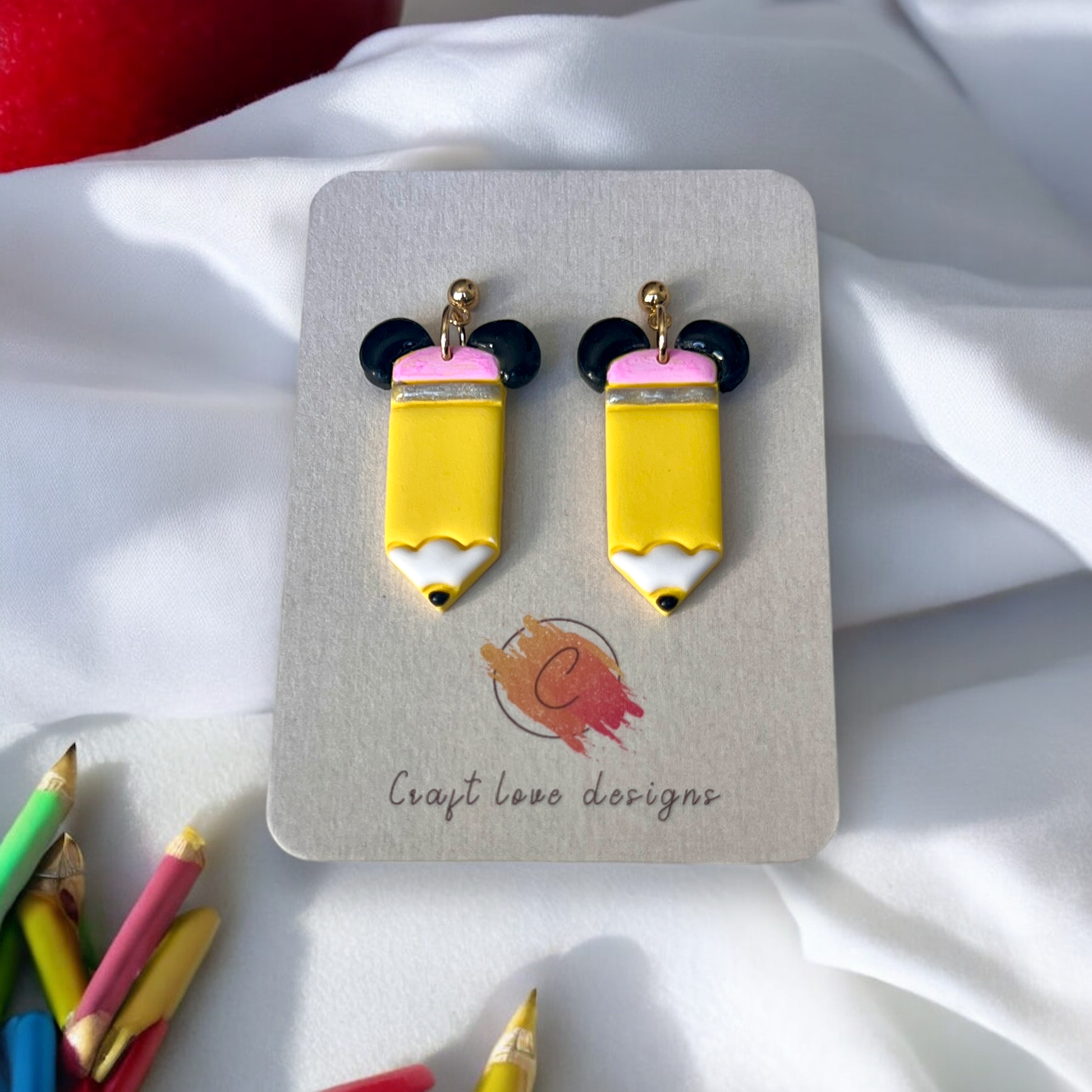 Yellow pencil-shaped earrings with black ear details and gold studs displayed on a designer card, surrounded by colorful pencils on a soft backdrop.