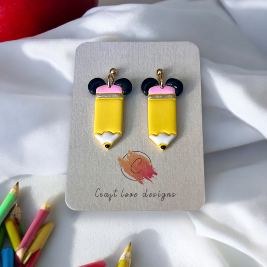 Yellow pencil-shaped earrings with black ear details and gold studs displayed on a designer card, surrounded by colorful pencils on a soft backdrop.