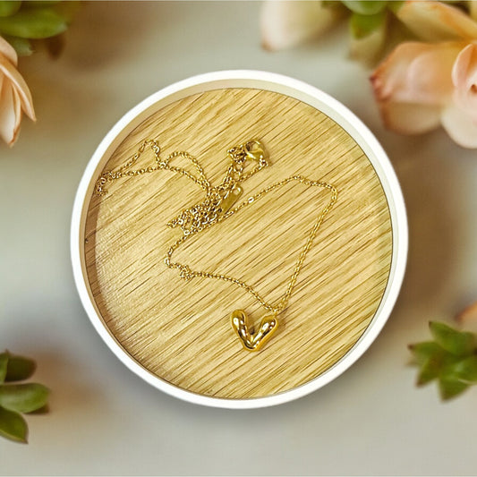 Elegant gold necklace featuring a petite puffy V heart pendant, delicately placed on a wooden surface surrounded by soft floral accents.