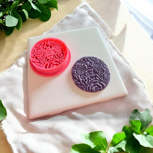 The pink cutter and finished purple clay piece showing a textured bee and floral design made with a 45mm polymer clay cutter