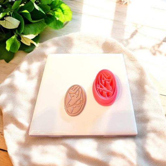 Red oval cutter next to nude-toned embossed feminine clay shape, displayed in natural light on a soft white board.