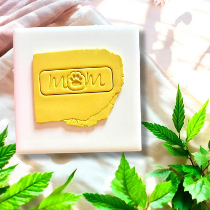Yellow clay slab with &quot;mom&quot; script and paw print tag impression, set on a white ceramic tile.