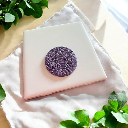 A round purple clay pendant featuring a bee surrounded by floral details, ready for necklace or keychain making