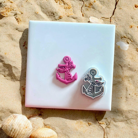 Pink mermaid tail anchor polymer clay charm alongside the white anchor cutter, displayed on white tile with beach sand.