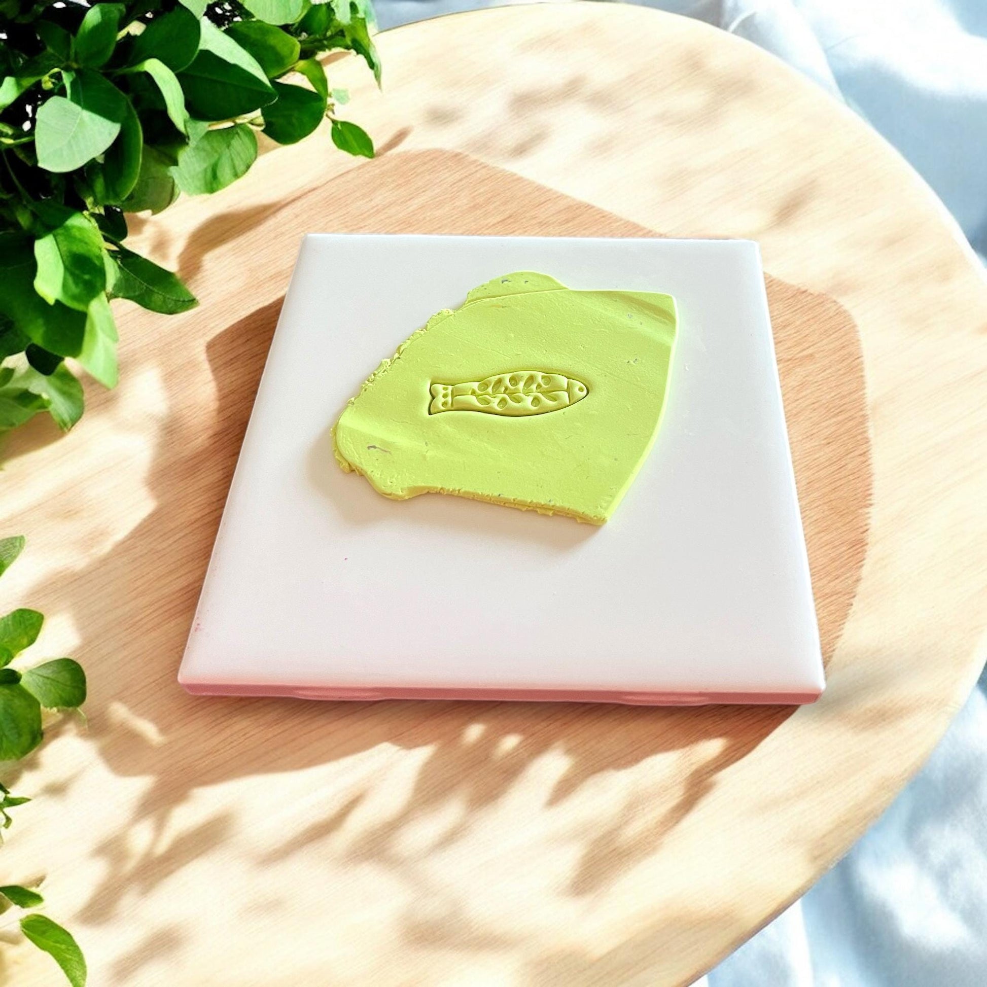 A green clay slab with a detailed floral fish design cutter impression under natural light on a white tile and fabric background