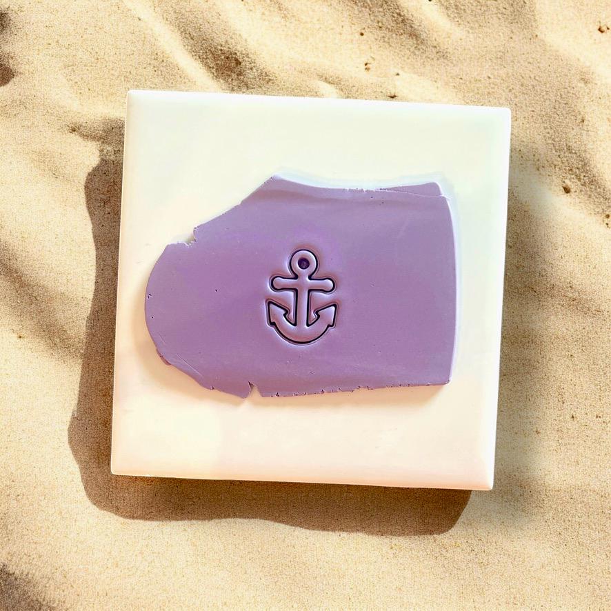 Anchor-shaped clay cutter used on purple polymer clay with a smooth impression, displayed on white tile with beach sand background.