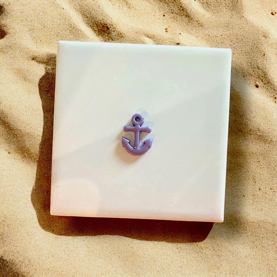 Small purple anchor charm made from polymer clay, displayed on a white ceramic tile with beach sand surroundings.