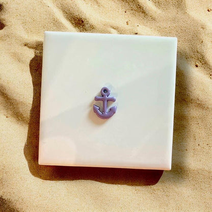 Small purple anchor charm made from polymer clay, displayed on a white ceramic tile with beach sand surroundings.