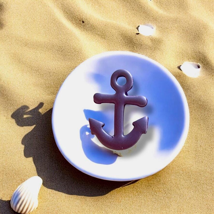 Large purple anchor charm made from polymer clay, set on a white plate with beach sand and seashell accents, perfect for nautical-themed jewelry.