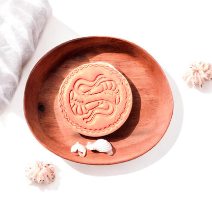 Finished shrimp clay design on wood tray surrounded by natural shells and light linen fabric.