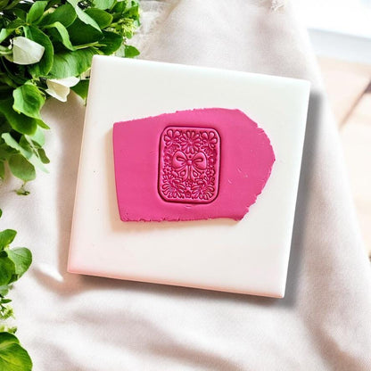 Coquette bow and floral motif clay imprint on pink polymer clay, displayed on white tile with greenery accents.