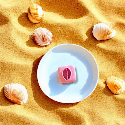 Pink cowrie shell cutter displayed on a white plate with beach sand and scattered seashells, perfect for summer-themed clay creations.
