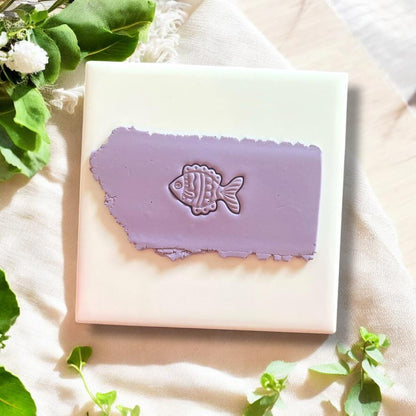 Detailed fish clay imprint on purple polymer clay, displayed on a white ceramic tile with fresh greenery accents.