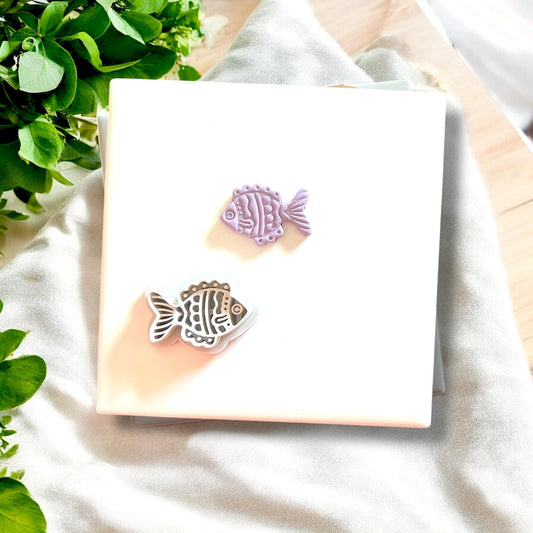 Polymer clay fish charm alongside its matching cutter, displayed on a white tile with fresh greenery.