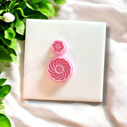 Pink polymer clay cutters in swirl shell designs, displayed on a white tile with green leaves.