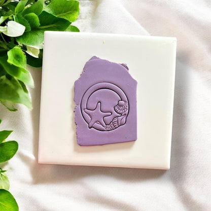 Purple polymer clay slab stamped with seashell circle cutter showing starfish and shell details on a white tile