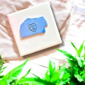 Turkish evil eye heart charm impression in blue polymer clay on a ceramic tile with fabric and green foliage.