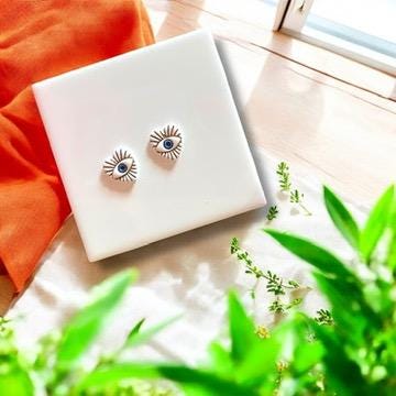 A pair of finished polymer clay evil eye heart earrings displayed on a tile with morning sunlight and greenery.