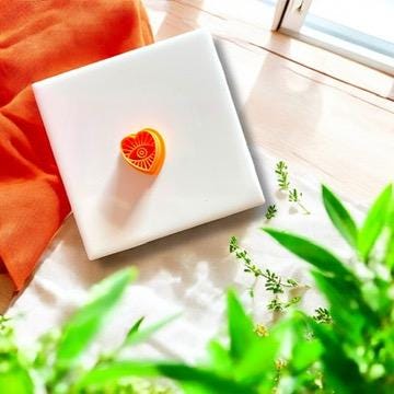 Turkish evil eye heart charm in orange clay shown with bold color contrast and greenery.
