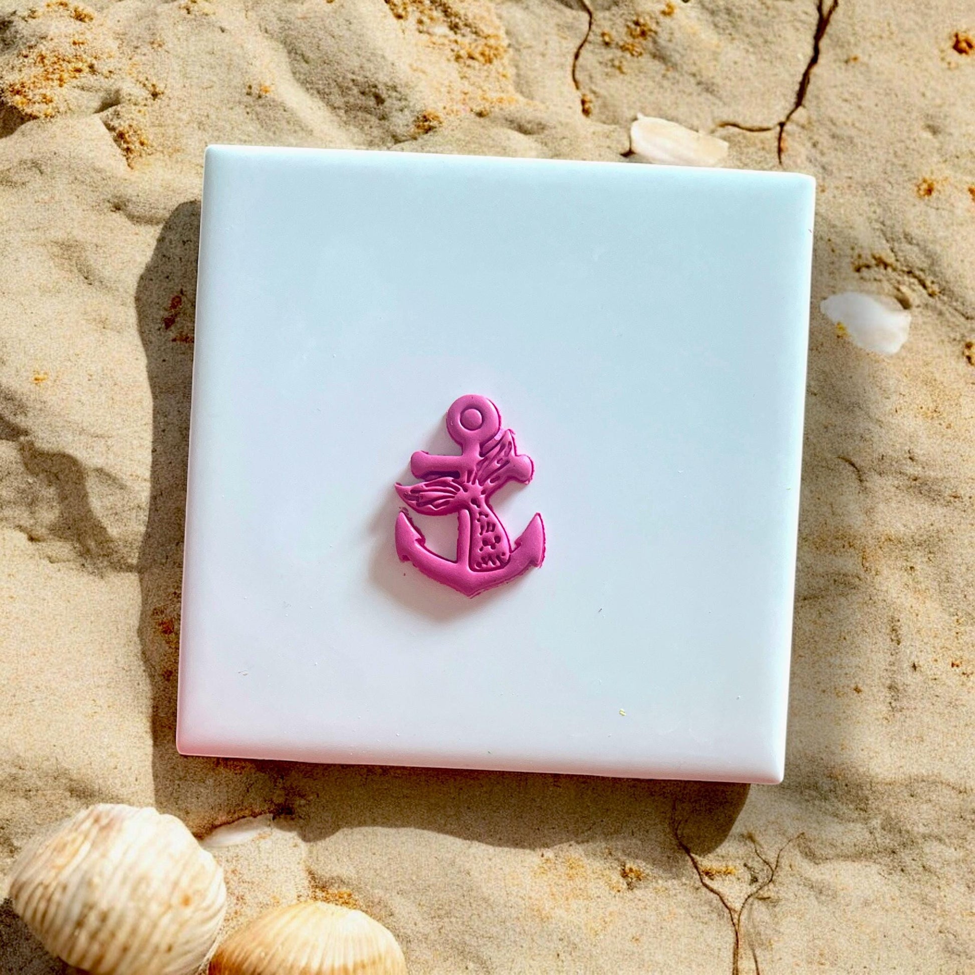 Finished polymer clay anchor with mermaid tail design on white tile with seashells and sand.